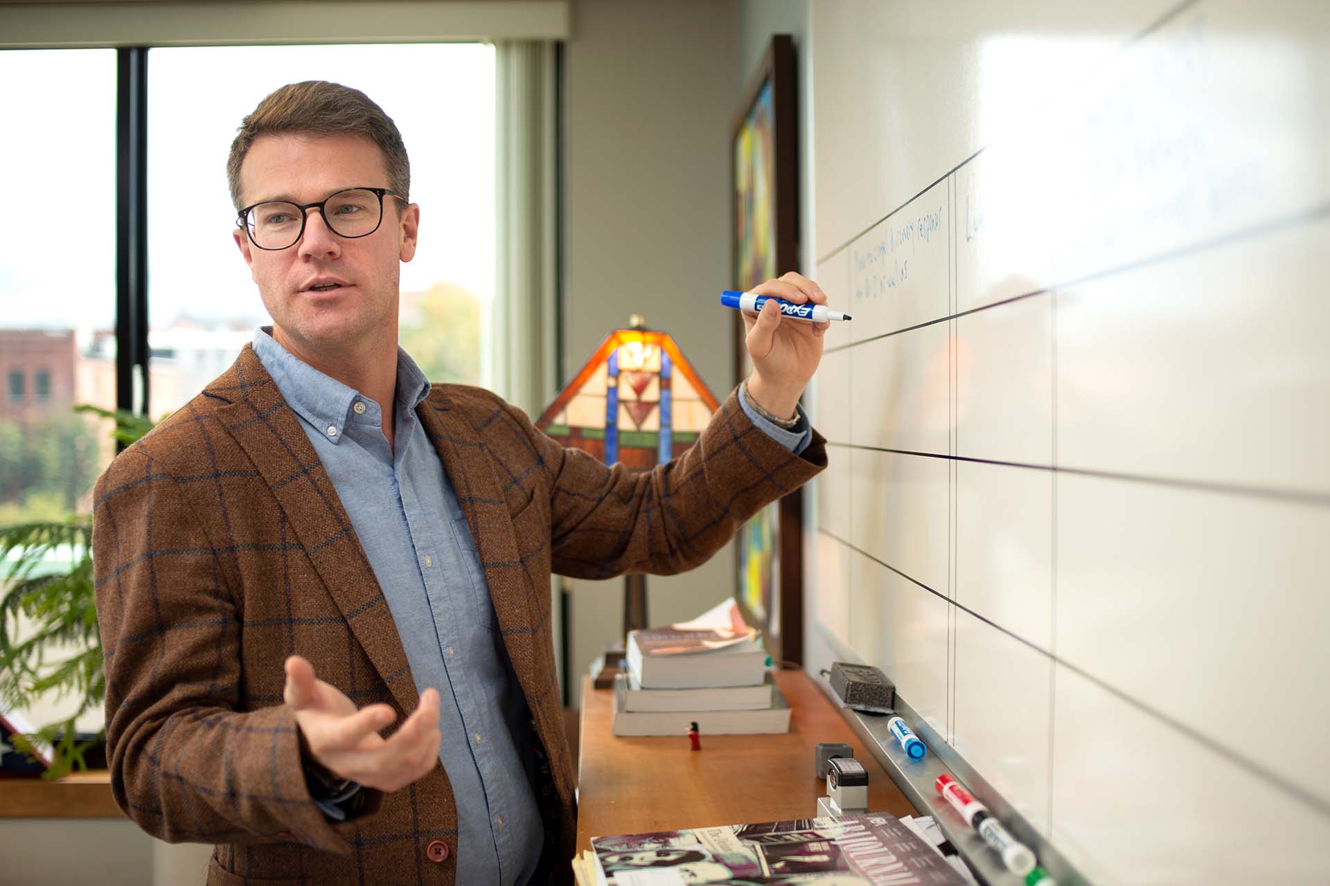 person in glasses and blazer talking while writing on white board