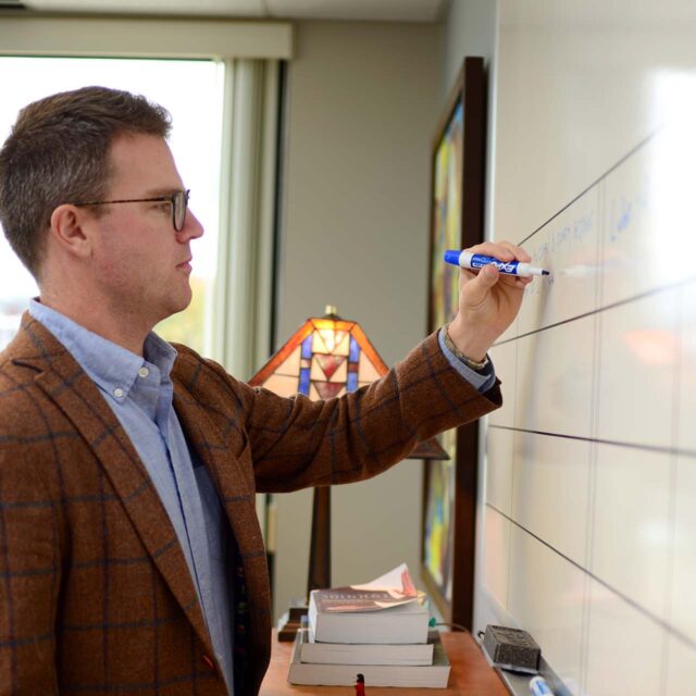 person with glasses and blazer writing on whiteboard on wall