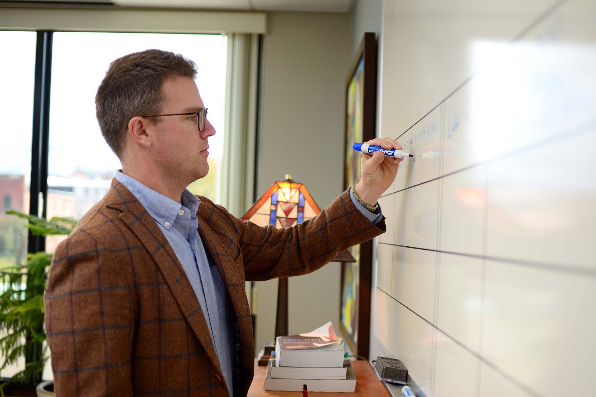 person with glasses and blazer writing on whiteboard on wall